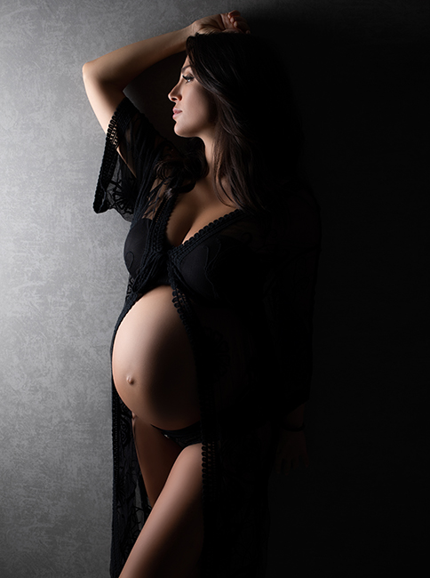 Schwangerschaftsfotografin Düsseldorf: Schwangere steht mit dem Rücken zur Wand in schwarzem Schwangerschaftskleid. Das Kleid öffnet sich über ihren Bauch.
