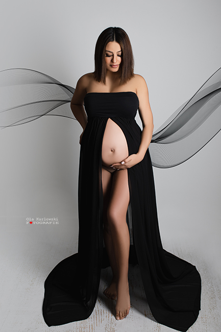 Schwangerschaftsfotografie Düsseldorf. Schwangere in Schwangerschaftskleid steht einem Engel gleich. Sie hält ihren Babybauch mit der linken Hand. Im Hintergrund schweben leichte Stoffbahnen. Das zweigeteilte schwarze Babybauchkleid gibt den Blick auf ihren nackten Babybauch frei.  