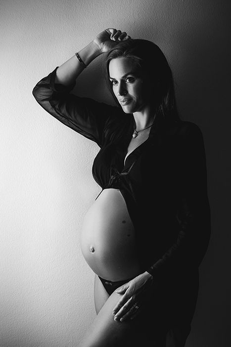 Schwangerschaftsfotografin Düsseldorf. Schwangere lehnt an Wand. Ihr Babybauch ist halb im Profil nackt zu sehen. Gekleidet ist sie mit einem schwarzem Schwangerschaftskleid. 