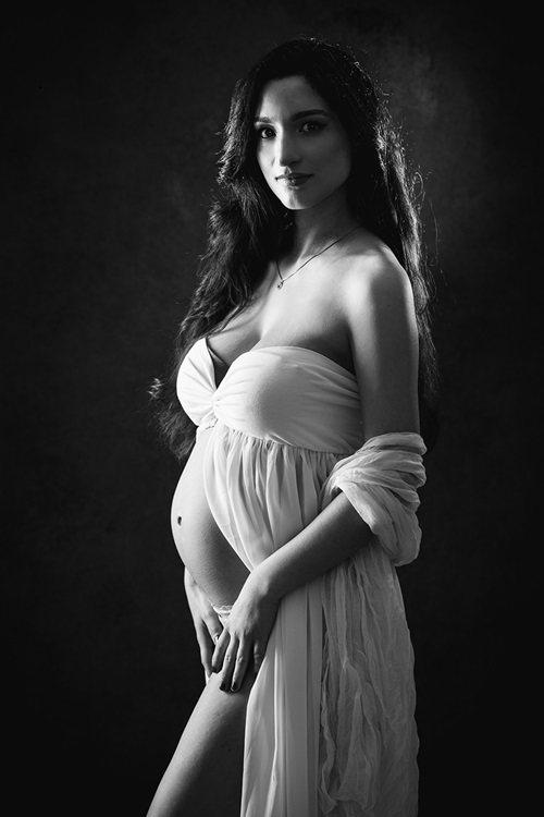 Schwangerschaftsfotografien Düsseldorf. Dasselbe Bild. Einmal in bunt, einmal schwarzweiß. Schwangere seitlich, mit leichtem Schwangerschaftskleid bekleidet, in die Kamera schauend.